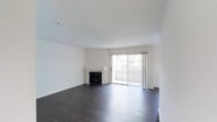 This single-floor space offers a clean and functional layout designed to maximize comfort and usability. The living area serves as the centerpiece of the space, finished with rich, dark hardwood flooring that contrasts elegantly against the crisp white walls and smooth ceiling. A classic fireplace with a simple white mantel anchors one side of the room, providing a focal point and a sense of warmth. On the opposite end, large sliding glass doors flood the room with natural light and provide direct access to an outdoor balcony, framing views of the greenery beyond. The sheer vertical drapes flanking the doors add a soft, airy touch to the overall aesthetic. The open layout of the living area creates a generous sense of space, offering versatile possibilities for furniture arrangement and interior styling. The neutral color palette throughout — from the white walls to the bright ceiling — enhances the sense of openness and makes the space feel welcoming and adaptable. Overall, this space combines functional design with understated elegance, making it a comfortable and inviting environment suited to a variety of lifestyles.