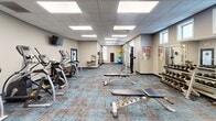 Located at 2105 S Silverthorne Ave in Sioux Falls, South Dakota, this single-floor fitness center is thoughtfully designed to support a comprehensive workout experience. The space features a modern, open layout with a drop-panel ceiling outfitted with bright recessed lighting panels that evenly illuminate the entire facility.

The main exercise area is well-equipped with multiple treadmills and elliptical cross-trainers arranged in rows, each positioned beneath wall-mounted flat-screen televisions for entertainment during cardio sessions. Adjustable weight benches and a dumbbell rack with a full range of free weights provide ample strength training options, while a cable/functional training machine adds versatility to the equipment lineup.

A dedicated flexibility and functional fitness zone is set apart from the main floor, featuring stability balls in multiple sizes, a wall-mounted exercise mat, resistance bands, and large windows fitted with roller shades that allow for natural light control. This area offers a quieter environment suited for stretching, balance training, or rehabilitation exercises.

Throughout the facility, a textured blue and tan patterned carpet tiles the floor, adding visual warmth and comfort underfoot. Neutral-toned walls, ample mirror placement, and clearly posted exercise reference charts contribute to a clean, organized, and user-friendly environment. The space is well-suited for a broad range of fitness activities and ability levels.