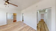 This renovated single-floor unit showcases a clean, modern aesthetic with light-toned hardwood flooring extending seamlessly throughout every room, creating a warm and cohesive atmosphere. The galley-style kitchen is well-appointed with dual-toned gray shaker cabinets lining both walls, crisp white countertops, stainless steel appliances including a gas range and refrigerator, and a range hood — offering an efficient and stylish cooking environment. The bathroom stands out with floor-to-ceiling marble-look tile in rich gray tones, a soaking tub with overhead shower, chrome fixtures, and a frosted window that allows natural light while maintaining privacy. The primary bedroom is generously proportioned, featuring mirrored sliding closet doors, a ceiling fan with light fixture, multiple windows with urban views, and a wall-mounted air conditioning unit. Additional rooms throughout the unit are bright and versatile, each with clean white walls, natural light from windows, and consistent hardwood flooring. A hallway connects the various spaces, providing a logical and functional layout. The overall ambiance is fresh, modern, and move-in ready, reflecting careful attention to detail in its renovation.