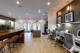This single-floor communal space is thoughtfully designed to serve multiple functions within one open, flowing layout. Light wood-look flooring runs throughout, creating a warm, cohesive foundation that ties each zone together seamlessly.

The lounge area anchors one end of the space, furnished with plush teal velvet sofas and accent chairs arranged around a round coffee table. Large windows dressed with natural woven roman shades and linen drapes allow generous natural light to fill the room, while a wall-mounted flat-screen television adds a media-ready element.

At the heart of the space, a substantial wood-topped communal bar table with woven rattan barstools serves as a casual gathering point. An adjacent dining area features a round table with sleek dark chairs, ideal for small group meetings or meals.

The co-working zone is equipped with a built-in desk station featuring multiple computer workstations and ergonomic seating. Nearby, a fully appointed kitchen includes dark cabinetry, stainless steel appliances, a full-size refrigerator, microwave, and a central island with bar seating. Accent walls in charcoal and warm brown, recessed lighting, and curated artwork throughout lend the space a polished, modern ambiance.