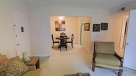 This single-floor residence showcases a thoughtfully designed open layout that seamlessly connects the living and dining areas, creating a cohesive and functional flow. The living room, visible in the foreground, features plush wall-to-wall carpeting in a neutral beige tone that extends throughout the shared space, contributing to a warm and inviting atmosphere.

A wide, open archway frames the transition into the dining area, where a round dark-wood dining table with four matching cross-back chairs serves as the focal point. A pendant light fixture overhead provides soft, direct illumination over the dining set. Wall art in earth tones adds visual interest to the dining space, complemented by decorative wall-mounted pieces on the adjacent wall.

A doorway visible beyond the dining area suggests access to additional rooms, indicating practical connectivity throughout the home. The overall color palette relies on soft whites and beiges, lending the space a calm, neutral character. The single-floor layout ensures easy accessibility and a straightforward, efficient use of space throughout.