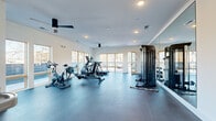 Located at 720 E 36th St in Charlotte, NC, this fitness center offers a thoughtfully designed workout environment within a compact yet efficiently utilized single-floor layout. The space is defined by its clean, modern aesthetic, featuring crisp white walls, recessed lighting, and durable dark rubberized flooring that provides both safety and comfort underfoot. Ceiling fans contribute to air circulation throughout the room, enhancing the overall workout experience. Large windows line multiple walls, inviting abundant natural light and offering views of the surrounding exterior, while glass-panel entry doors add a sleek, open feel to the space. The fitness center is well-appointed with a variety of equipment, including cycling bikes, an elliptical trainer, a treadmill, and a multi-station cable machine by Inspire, along with a free weight rack. A full-length mirror panel is positioned alongside the strength equipment, supporting proper form during workouts. A built-in water fountain and a small open storage cubby unit provide added convenience. An additional door leads to what appears to be an outdoor area, further extending accessibility. The overall ambiance is bright, functional, and inviting, making it a practical and well-maintained facility for residents.