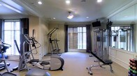 This dedicated fitness center is thoughtfully designed to provide a comprehensive workout environment within a single, open-plan floor. The space is outfitted with a range of cardio equipment, including elliptical trainers and a stair climber, positioned along one side of the room for easy access. On the opposite side, a cable weight machine and a weight bench are set against a floor-to-ceiling mirrored wall, allowing users to monitor their form during strength training sessions. A wall-mounted flat-screen television adds convenience for entertainment during workouts. The room benefits from natural light streaming through a large window dressed with wooden blinds and elegant floor-length curtains, lending a welcoming ambiance to the space. Recessed ceiling lighting, complemented by a central ceiling fan, ensures the area remains well-lit and comfortable at any time of day. The light-toned carpeted flooring and neutral wall colors contribute to a clean, focused atmosphere. Crown molding details along the ceiling add a refined architectural touch, elevating the overall aesthetic of the fitness space.