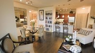 This single-floor residence showcases a thoughtfully designed open-concept layout that unifies the living, dining, and kitchen areas into one cohesive and inviting space. Rich, dark hardwood flooring flows continuously throughout, anchoring the warm, sophisticated aesthetic that defines the interior.

The dining area features a round glass-top table surrounded by upholstered chairs in a neutral tone, complemented by a statement mirror on the wall that enhances the sense of depth and light. An integrated built-in shelving unit serves as a functional and decorative divider between the dining and kitchen zones, offering display space for books and decorative accents.

The kitchen is appointed with warm wood cabinetry, granite or stone countertops, and stainless steel appliances. A raised breakfast bar with cross-back bar stools provides casual seating and extends the kitchen's functionality. Pendant lighting and a track lighting system illuminate both the kitchen and surrounding areas, creating a well-lit, layered lighting scheme.

Track lighting along the ceiling and recessed fixtures contribute to the overall warm ambiance, making the space feel both functional and refined throughout.