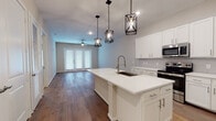 This single-floor residence offers a thoughtfully designed open-concept layout that seamlessly connects the kitchen and living areas. The kitchen serves as the centerpiece of the home, showcasing a large island with a white quartz countertop, an undermount sink with a pull-down faucet, and a full suite of stainless steel appliances including a refrigerator, range, microwave, and dishwasher. Crisp white shaker-style cabinetry lines the walls, while three geometric black pendant lights overhead add a striking industrial-modern touch. Rich hardwood flooring flows throughout the main living area, which is anchored by a ceiling fan and French doors that invite natural light and provide outdoor access. The bedrooms feature plush carpeting and ceiling fans, offering a comfortable, quieter retreat. The bathrooms are finished with neutral tile flooring and provide functional, clean aesthetics — one features a wide vanity with ample cabinet storage beneath a large mirror with bright overhead lighting, while another includes a full soaking tub and shower combo with elegantly tiled surrounds. Overall, the space combines contemporary finishes with a warm, livable atmosphere suited for everyday comfort.