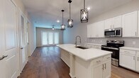 This single-floor residence offers a thoughtfully designed open-concept layout that seamlessly connects the kitchen and living areas. The kitchen serves as the centerpiece of the home, showcasing a large island with a white quartz countertop, an undermount sink with a pull-down faucet, and a full suite of stainless steel appliances including a refrigerator, range, microwave, and dishwasher. Crisp white shaker-style cabinetry lines the walls, while three geometric black pendant lights overhead add a striking industrial-modern touch. Rich hardwood flooring flows throughout the main living area, which is anchored by a ceiling fan and French doors that invite natural light and provide outdoor access. The bedrooms feature plush carpeting and ceiling fans, offering a comfortable, quieter retreat. The bathrooms are finished with neutral tile flooring and provide functional, clean aesthetics — one features a wide vanity with ample cabinet storage beneath a large mirror with bright overhead lighting, while another includes a full soaking tub and shower combo with elegantly tiled surrounds. Overall, the space combines contemporary finishes with a warm, livable atmosphere suited for everyday comfort.