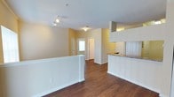 This single-floor space showcases an open-concept design that seamlessly connects the main living area to the kitchen. The flooring throughout is a warm, medium-toned hardwood-style material that adds a cohesive and inviting feel to the space. A textured ceiling runs throughout, complemented by a combination of track lighting and ceiling-mounted fixtures that ensure the interior is well-illuminated.

The living area is defined by a low partial-wall divider, creating a subtle separation while maintaining the open flow of the layout. Walls are painted in a soft, neutral yellow-beige tone that contributes to a warm and welcoming ambiance.

The kitchen is positioned to one side and features white cabinetry, granite-style countertops, and a raised breakfast bar that overlooks the living space. An overhead cabinet structure adds additional storage and visual definition to the kitchen zone.

Toward the rear of the space, a doorway framed by natural light hints at access to additional rooms or an exterior entry. The overall layout is functional and efficient, with clean lines, neutral finishes, and a bright character throughout.