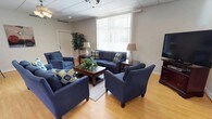 This single-floor space functions as a welcoming common lounge or sitting area, designed to encourage relaxed gathering and conversation. The room is furnished with a coordinated navy blue seating arrangement, including a full sofa, loveseat, and two accent armchairs, all accented with blue and yellow patterned throw pillows that add a pop of color to the neutral palette. A dark wood coffee table anchors the central seating group, accompanied by matching end tables, each topped with classic white table lamps that contribute warm, ambient lighting.

The flooring consists of light hardwood-style planks that extend throughout the space, lending warmth and a clean, open feel. A large window fitted with vertical blinds occupies a prominent wall, allowing generous natural light to enter and providing views of the greenery outside. A tall decorative indoor plant in the corner adds a natural, refreshing element to the room.

The ceiling features a drop-tile design with recessed lighting and a ceiling fan, ensuring the space remains comfortable year-round. A flat-screen television mounted near the wall rounds out the amenities, making this a functional, well-appointed space suited for socializing or quiet relaxation.