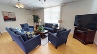This single-floor space functions as a welcoming common lounge or sitting area, designed to encourage relaxed gathering and conversation. The room is furnished with a coordinated navy blue seating arrangement, including a full sofa, loveseat, and two accent armchairs, all accented with blue and yellow patterned throw pillows that add a pop of color to the neutral palette. A dark wood coffee table anchors the central seating group, accompanied by matching end tables, each topped with classic white table lamps that contribute warm, ambient lighting.

The flooring consists of light hardwood-style planks that extend throughout the space, lending warmth and a clean, open feel. A large window fitted with vertical blinds occupies a prominent wall, allowing generous natural light to enter and providing views of the greenery outside. A tall decorative indoor plant in the corner adds a natural, refreshing element to the room.

The ceiling features a drop-tile design with recessed lighting and a ceiling fan, ensuring the space remains comfortable year-round. A flat-screen television mounted near the wall rounds out the amenities, making this a functional, well-appointed space suited for socializing or quiet relaxation.