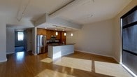 This single-floor space showcases a thoughtfully designed open-concept layout that seamlessly blends the kitchen and living areas into one cohesive, light-filled environment. The kitchen is a standout feature, equipped with stainless steel appliances including a full-size refrigerator and built-in microwave, complemented by rich warm-toned wood cabinetry that adds depth and character. A central island with a dark countertop serves as both a functional workspace and a natural focal point, anchoring the kitchen within the open floor plan. Pendant lighting hangs above the island, providing focused illumination while contributing to the space's modern aesthetic. Track lighting runs along the ceiling, offering flexible, adjustable light throughout. The living area extends generously from the kitchen, enhanced by wide-plank light hardwood flooring that runs continuously throughout the space, reflecting natural sunlight and creating a warm, inviting ambiance. The high ceilings, accentuated by exposed structural beams, lend an airy, loft-like quality to the interior. The neutral gray walls provide a clean, versatile backdrop, making the space adaptable to a variety of uses and design preferences.