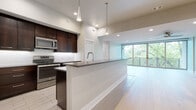 This single-floor residence showcases a contemporary design with a thoughtfully planned open layout. The kitchen is a standout feature, equipped with stainless steel appliances including a refrigerator, range, and microwave, all set against rich dark-wood cabinetry and a classic white subway tile backsplash. A large peninsula with a dark countertop and an integrated sink provides generous workspace and serves as a natural divider between the kitchen and the living area. Sleek pendant lights hang above, adding a modern touch.

The living area is bright and airy, benefiting from floor-to-ceiling glass doors that frame lush outdoor greenery and open to a balcony with a horizontal railing. Light wood-style flooring runs throughout the main living spaces, lending warmth and continuity. Recessed lighting and a ceiling fan ensure comfortable illumination throughout.

The bedroom is generously proportioned, featuring the same light hardwood flooring and soft neutral walls. It connects to a walk-in closet outfitted with built-in shelving and hanging rods. The bathroom maintains the modern aesthetic with a dark-wood vanity, white countertop, undermount sink, large mirror, and neutral tile flooring, creating a clean, polished finish throughout.