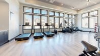 This modern fitness center offers a thoughtfully designed, single-floor workout space that provides ample room for a full range of exercise activities. The layout is open and well-organized, with light hardwood flooring throughout that contributes to the clean, contemporary aesthetic.

The cardio zone is positioned along a dramatic floor-to-ceiling window wall, flooding the area with abundant natural light and offering views of the surrounding community. This section is equipped with multiple treadmills, elliptical trainers, a stair climber, and a rowing machine, providing a comprehensive selection of cardiovascular options.

The strength training area features a robust collection of equipment, including cable machines, adjustable benches, and dedicated free weight stations with a full rack of dumbbells. Floor-to-ceiling mirrors line the walls in this zone, enhancing the sense of space and allowing users to monitor their form during workouts.

The high ceilings with a tray detail and a large ceiling fan contribute to an airy, spacious ambiance throughout the facility. Built-in shelving and cabinetry along one wall provide practical storage, while a wall-mounted television adds an entertainment element. The space is well-ventilated with visible HVAC ceiling vents, ensuring a comfortable environment for all fitness activities.
