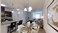 This single-floor residence offers a thoughtfully designed open-concept layout that seamlessly connects the main living areas, creating a fluid and welcoming environment. The dining area is a standout feature, anchored by an elegant multi-light chandelier with brushed nickel finishes that casts a warm glow over a glass-top dining table surrounded by contrasting black and white chairs. Hardwood-style flooring runs throughout, adding warmth and visual continuity to the space. Adjacent to the dining area, the kitchen is partially visible and features pendant lighting, granite-style countertops, and white cabinetry, offering both style and functionality. The living room extends naturally from the dining space, bathed in natural light from a large window and cooled by a ceiling fan overhead. Neutral gray walls and light-toned ceilings keep the atmosphere bright and airy, while the open layout enhances the overall sense of spaciousness. The combination of modern lighting fixtures, clean architectural lines, and a cohesive neutral color palette gives this residence a polished, contemporary feel that is both functional and visually appealing.