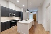 Located at 1557 Logan Street in Denver's Capitol Hill neighborhood, this modern 1-bedroom, 1-bathroom unit offers a well-designed single-floor layout with contemporary finishes throughout. The open-concept kitchen serves as the centerpiece of the space, featuring bold dark cabinetry that extends to the ceiling, a large white quartz island with an undermount sink and gold-toned faucet, a tile backsplash, and a full suite of stainless steel appliances including a gas range, built-in microwave, and French door refrigerator. Sleek pendant lights hang above the island, adding a modern touch. Warm-toned hardwood flooring runs throughout the entire unit, creating a cohesive and inviting aesthetic. The entry area benefits from natural light through a frosted glass window and features a natural wood front door. The bedroom is a comfortable size, offering a large window with diffused natural light, clean white walls, and ample room for furnishings. The bathroom includes black hardware accents, a glass-enclosed shower, and continues the unit's consistent modern design language. Additional features include a dedicated closet and an in-unit laundry area, providing practical convenience within a compact, efficiently laid-out space.