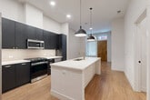Located at 1557 Logan Street in Denver's Capitol Hill neighborhood, this modern 1-bedroom, 1-bathroom unit offers a well-designed single-floor layout with contemporary finishes throughout. The open-concept kitchen serves as the centerpiece of the space, featuring bold dark cabinetry that extends to the ceiling, a large white quartz island with an undermount sink and gold-toned faucet, a tile backsplash, and a full suite of stainless steel appliances including a gas range, built-in microwave, and French door refrigerator. Sleek pendant lights hang above the island, adding a modern touch. Warm-toned hardwood flooring runs throughout the entire unit, creating a cohesive and inviting aesthetic. The entry area benefits from natural light through a frosted glass window and features a natural wood front door. The bedroom is a comfortable size, offering a large window with diffused natural light, clean white walls, and ample room for furnishings. The bathroom includes black hardware accents, a glass-enclosed shower, and continues the unit's consistent modern design language. Additional features include a dedicated closet and an in-unit laundry area, providing practical convenience within a compact, efficiently laid-out space.