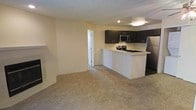 This single-floor space showcases an open-concept layout that seamlessly connects the living area to the kitchen. The carpeted living area offers a comfortable, neutral backdrop with warm beige tones and smooth walls painted in a soft cream color, contributing to an airy and welcoming ambiance throughout.

The kitchen is a standout feature, equipped with dark espresso-toned cabinetry that provides a striking contrast against the light-colored countertops and walls. Stainless steel appliances, including a refrigerator, microwave, and range, add a modern and functional touch. A peninsula-style breakfast bar extends from the kitchen, creating a natural divide between the cooking and living spaces while also providing additional countertop space for dining or meal preparation.

A doorway visible from the main living area leads to an adjacent room or hallway, suggesting further utility space beyond what is immediately visible. Overhead lighting illuminates the kitchen area effectively. The neutral color palette, open layout, and combination of carpet and smooth surfaces make this a practical and versatile space well-suited for everyday living.