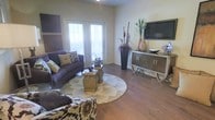 This single-floor space offers a thoughtfully designed living area that blends comfort and contemporary style. The living room serves as the focal point, showcasing rich hardwood flooring that runs throughout and anchors the space with warmth. A circular area rug defines the seating arrangement, which features a plush purple sofa accented with decorative pillows and complementary patterned accent chairs, creating an inviting and cohesive aesthetic.

A pair of elegant French doors floods the room with natural light, opening the space visually and connecting the interior to the outside. A wall-mounted flat-screen television sits above a sleek, modern media console styled with decorative vases and botanical accents, adding a refined touch to the entertainment area.

Tall decorative plants and curated accessories, including sculptural floor vases and tabletop arrangements, bring organic texture and visual interest to the room. A distinctive pendant light fixture hangs from the ceiling, adding a subtle architectural detail overhead.

The overall ambiance is polished yet approachable, with a neutral color palette warmed by earthy tones and deep purples. The layout feels open and well-proportioned, making it both functional for everyday living and visually impressive for entertaining.