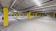 This single-level underground parking facility offers a well-organized and efficiently designed space for vehicle storage. The garage is characterized by its bold yellow structural columns, which are evenly distributed throughout the floor and serve as both load-bearing supports and high-visibility safety markers. The smooth, light-colored concrete floor is clearly marked with numbered parking stalls, allowing for easy navigation and space identification.

The ceiling features exposed concrete construction with a network of overhead pipes and conduit running throughout, providing a functional industrial aesthetic. Rows of fluorescent strip lighting illuminate the entire level uniformly, ensuring excellent visibility across all areas of the garage.

A notable feature of this facility is the inclusion of dedicated electric vehicle charging stations, equipped with charging hardware and clearly designated stalls, reflecting a forward-thinking approach to modern parking infrastructure. Along the perimeter, secure wire-mesh storage cages provide additional utility for residents or tenants.

The facility's open-plan layout offers generous circulation space between rows of stalls, accommodating easy vehicle maneuvering. The overall design prioritizes practicality, safety, and efficiency, making it a well-appointed underground parking solution suited to a contemporary residential or commercial building.