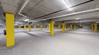 This single-level underground parking facility offers a well-organized and efficiently designed space for vehicle storage. The garage is characterized by its bold yellow structural columns, which are evenly distributed throughout the floor and serve as both load-bearing supports and high-visibility safety markers. The smooth, light-colored concrete floor is clearly marked with numbered parking stalls, allowing for easy navigation and space identification.

The ceiling features exposed concrete construction with a network of overhead pipes and conduit running throughout, providing a functional industrial aesthetic. Rows of fluorescent strip lighting illuminate the entire level uniformly, ensuring excellent visibility across all areas of the garage.

A notable feature of this facility is the inclusion of dedicated electric vehicle charging stations, equipped with charging hardware and clearly designated stalls, reflecting a forward-thinking approach to modern parking infrastructure. Along the perimeter, secure wire-mesh storage cages provide additional utility for residents or tenants.

The facility's open-plan layout offers generous circulation space between rows of stalls, accommodating easy vehicle maneuvering. The overall design prioritizes practicality, safety, and efficiency, making it a well-appointed underground parking solution suited to a contemporary residential or commercial building.