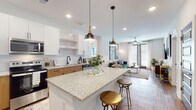 This well-appointed single-floor residence at 1290 Old Peachtree Rd NW in Duluth, Georgia, offers a thoughtfully designed open-concept layout with modern finishes throughout. The kitchen serves as the heart of the home, showcasing granite countertops, a large central island with seating, two-tone cabinetry combining crisp white upper cabinets with warm wood lower cabinets, and a classic subway tile backsplash. Stainless steel appliances, including a french door refrigerator, range, microwave, and dishwasher, complete this functional and stylish culinary space. Pendant lighting above the island adds a refined touch. The kitchen flows seamlessly into the living area, which features warm hardwood-style flooring, large windows with blinds, access to an outdoor area via glass doors, and a ceiling fan that enhances comfort. The bedrooms are carpeted and bright, each benefiting from generous natural light through wide windows dressed with layered blinds and curtains. The primary bathroom includes a granite-topped wood vanity and a glass-enclosed walk-in shower, with direct access to a walk-in closet. A secondary bathroom mirrors the same refined aesthetic with a matching vanity and tub-shower combination. The overall ambiance is modern, clean, and cohesive throughout.
