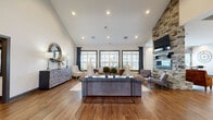 Located in Smyrna, Tennessee, this single-floor community space offers an open-concept layout that seamlessly connects the living room, dining area, and kitchen. The living room is a standout, showcasing soaring vaulted ceilings, rich hardwood-style flooring, and a dramatic floor-to-ceiling stacked stone fireplace with a wall-mounted television above. A bank of large, dark-framed windows floods the space with abundant natural light and provides views of the surrounding community. Recessed lighting complements the natural light throughout.

The seating area is expansive, accommodating multiple conversation zones, while a separate seating nook near the windows offers a bright, relaxed retreat. The dining area features high-top tables and chairs positioned alongside large windows dressed with decorative patterned valances, offering additional community views.

The kitchen is clean and functional, outfitted with dark cabinetry, crisp white countertops, a stainless steel sink, and built-in appliances. A central island adds prep space and versatility. Consistent hardwood-style flooring ties the entire space together, creating a cohesive, welcoming atmosphere well-suited for community gatherings and events.