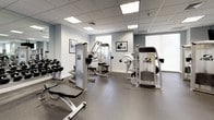 This fitness center is a thoughtfully designed, single-floor facility outfitted with a comprehensive selection of professional-grade exercise equipment. The space is anchored by a row of Cybex treadmills and elliptical machines, each equipped with individual monitors for entertainment during cardio sessions. A dedicated strength training area features multiple Cybex weight machines, a dumbbell rack with a full range of free weights, and adjustable benches, providing ample options for resistance training. Floor-to-ceiling mirrors line portions of the walls, enhancing the sense of openness and allowing users to monitor their form. The flooring throughout is durable rubberized material, offering both safety and comfort underfoot. Overhead, a recessed drop ceiling with flush panel lighting ensures the space is consistently and evenly illuminated. Light grey walls and a clean, neutral color palette contribute to a modern, professional atmosphere. Framed fitness-themed artwork is displayed on the walls, adding a motivational accent to the environment. A water fountain is conveniently located within the space. Glass doors at one end open directly to an outdoor area, allowing for natural light and easy access. The overall design is functional and well-organized, catering to a full range of cardio and strength training needs.