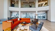 This contemporary leasing lobby makes a striking first impression with its dramatic double-height ceilings and an open, airy layout anchored by polished concrete flooring throughout. The main entrance welcomes visitors through floor-to-ceiling glass walls that flood the space with natural light and offer views of the surrounding exterior.

The central lounge area is a standout feature, defined by a bold L-shaped sectional sofa in deep orange velvet, complemented by charcoal upholstered armchairs and a richly patterned area rug. The seating arrangement is anchored beneath a sweeping upper-level mezzanine, edged with glass railings that add to the sense of vertical volume.

Lining the walls are large-scale, hand-illustrated murals depicting iconic city landmarks and architectural scenes, lending the lobby a strong sense of local identity and urban character. These murals serve as dramatic backdrops to both the lounge and a casual communal work table, crafted from live-edge wood with industrial-style metal legs and paired with leather-look chairs.

Adjacent to the main lobby, a glass-enclosed conference room provides a private meeting space, outfitted with a formal table and chairs. A separate room with a sliding barn door adds a warm, transitional touch. The overall design balances modern industrial elements with inviting furnishings, creating a welcoming and functional environment.