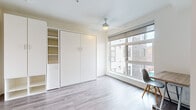 Located in Portland's vibrant NW district, this efficiently designed studio offers a smart, modern layout that makes excellent use of its compact footprint. The main living area features light gray hardwood-style flooring, crisp white walls, and a ceiling fan overhead. A large, multi-pane window floods the space with natural light while framing views of the surrounding neighborhood. Built-in white cabinetry with integrated open shelving provides generous storage, complemented by a fold-away murphy bed that maximizes the flexibility of the living area. A wall-mounted desk with a wood-top surface offers a dedicated workspace beside the window. The kitchen is thoughtfully equipped with stainless steel appliances, including a full-size refrigerator, dishwasher, and built-in microwave, alongside white upper cabinetry, light-toned lower drawers, a smooth cooktop, and a single-basin sink with a modern faucet. The bathroom includes a shower/tub combination with chrome fixtures and a grab bar, finished with wood-look vinyl flooring and clean white walls throughout. The overall aesthetic is clean, contemporary, and functional — well-suited for efficient urban living in the heart of Portland.