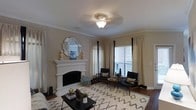 This single-story residence offers a thoughtfully designed layout within a comfortable footprint. The living room serves as the centerpiece of the home, showcasing a striking white fireplace with a detailed mantel surround that anchors the space with classic elegance. Above the mantel, an octagonal mirrored accent piece enhances the room's sense of depth and light. Crown molding runs along the ceiling perimeter, adding a refined architectural touch to the warm, neutral-toned walls. A flush-mount ceiling fixture with a decorative glass shade provides ambient lighting throughout the space. Large windows dressed with floor-length sheer and blackout panel curtains allow natural light to filter into the room while maintaining privacy. Dark hardwood flooring grounds the space and contrasts beautifully with a bold geometric area rug in black and white. Mid-century modern accent chairs and a sleek coffee table complete the living area's sophisticated aesthetic. The overall ambiance is warm, inviting, and polished, blending classic architectural details with contemporary furnishings for a balanced and livable space.