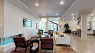 This well-appointed clubroom offers a sophisticated and inviting atmosphere designed for comfort and socializing. The space features an open-concept layout with distinct yet seamlessly connected lounge areas, each thoughtfully furnished to encourage both relaxed gatherings and casual conversation. A sleek wall-mounted flat-screen television is positioned above a fireplace framed by a textured white brick accent wall, adding warmth and visual interest to the main seating area. Rich, patterned area rugs anchor the furniture groupings, which include a plush white sofa accented with teal and gold throw pillows, deep red upholstered armchairs, and a dark wood table with coordinating seating. Overhead, a textured ceiling with recessed lighting and a central tray detail with ambient LED illumination adds architectural dimension to the room. A statement pendant light hangs above, complementing the layered lighting throughout. Large windows and glass doors at the far end draw in abundant natural light and provide views to an outdoor area, creating a bright and airy ambiance. Lush indoor plants enhance the space with a touch of greenery, while the overall design palette of neutral tones, warm woods, and bold accent colors conveys a polished yet approachable character.