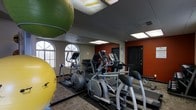 This fitness center is a fully equipped exercise facility designed to accommodate a variety of workout routines. The space features a selection of cardio machines, including ellipticals and treadmills, arranged to allow comfortable use and easy movement throughout the room. Large exercise balls in vibrant green and yellow add a versatile element for strength and balance training. The interior is finished with a drop ceiling fitted with recessed fluorescent lighting that ensures the space is well-lit throughout. Arched windows along one wall invite natural light, contributing to an open and energizing atmosphere. A bold terracotta-colored accent wall adds a dynamic visual contrast to the neutral tones of the rest of the space. Full-length mirrors line one wall, enhancing the sense of space while allowing users to monitor their form during workouts. The flooring features a durable, low-pile carpet suited to the demands of a fitness environment. A single-floor layout ensures all amenities are conveniently accessible, making this fitness center a practical and motivating space for daily exercise.