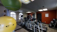 This fitness center is a fully equipped exercise facility designed to accommodate a variety of workout routines. The space features a selection of cardio machines, including ellipticals and treadmills, arranged to allow comfortable use and easy movement throughout the room. Large exercise balls in vibrant green and yellow add a versatile element for strength and balance training. The interior is finished with a drop ceiling fitted with recessed fluorescent lighting that ensures the space is well-lit throughout. Arched windows along one wall invite natural light, contributing to an open and energizing atmosphere. A bold terracotta-colored accent wall adds a dynamic visual contrast to the neutral tones of the rest of the space. Full-length mirrors line one wall, enhancing the sense of space while allowing users to monitor their form during workouts. The flooring features a durable, low-pile carpet suited to the demands of a fitness environment. A single-floor layout ensures all amenities are conveniently accessible, making this fitness center a practical and motivating space for daily exercise.