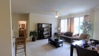 This single-floor residence offers a comfortable and thoughtfully laid-out living environment. The main living area is a welcoming space anchored by plush wall-to-wall carpeting and neutral-toned walls that create a clean, airy atmosphere. A ceiling fan with a light fixture is centrally positioned overhead, providing both functional airflow and ambient lighting. Large windows dressed with warm, draped curtains allow generous natural light to flood the room, enhancing its open and inviting feel. The living area accommodates a full seating arrangement centered around a dark wood coffee table, complemented by a matching bookcase and entertainment unit that add both storage and visual structure to the space. Lush indoor greenery adds a touch of nature and warmth to the room. Crown molding trims the ceiling, lending an added touch of architectural detail. An open sightline connects the living area to an adjacent dining space, where wooden chairs are visible, suggesting a seamlessly connected layout ideal for everyday living and entertaining. A hallway leading to a vanity-lit area hints at additional rooms beyond, completing a residence designed for both practicality and comfort.