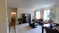 This single-floor residence offers a comfortable and thoughtfully laid-out living environment. The main living area is a welcoming space anchored by plush wall-to-wall carpeting and neutral-toned walls that create a clean, airy atmosphere. A ceiling fan with a light fixture is centrally positioned overhead, providing both functional airflow and ambient lighting. Large windows dressed with warm, draped curtains allow generous natural light to flood the room, enhancing its open and inviting feel. The living area accommodates a full seating arrangement centered around a dark wood coffee table, complemented by a matching bookcase and entertainment unit that add both storage and visual structure to the space. Lush indoor greenery adds a touch of nature and warmth to the room. Crown molding trims the ceiling, lending an added touch of architectural detail. An open sightline connects the living area to an adjacent dining space, where wooden chairs are visible, suggesting a seamlessly connected layout ideal for everyday living and entertaining. A hallway leading to a vanity-lit area hints at additional rooms beyond, completing a residence designed for both practicality and comfort.