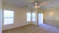 This single-floor space offers a welcoming, open living area characterized by vaulted ceilings that lend a sense of height and airiness. A ceiling fan with a built-in light fixture is centrally positioned, providing both comfort and ambient lighting. The room is finished with neutral-toned carpeting that extends throughout, contributing to a warm and understated aesthetic.

Natural light is a defining feature, with multiple windows positioned along two walls, including a corner arrangement that allows light to pour in from multiple directions. Vertical blinds on a sliding glass door provide access to the outdoors while offering privacy and light control when needed. The combination of windows and the door ensures the space remains bright throughout the day.

The walls are finished in a soft, light neutral tone, complementing the clean white trim and baseboards that frame the room. A textured ceiling adds subtle visual interest overhead. The layout is open and versatile, allowing for a variety of furniture arrangements. Overall, the space presents a clean, functional, and naturally lit environment suited for everyday living.