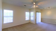 This single-floor space offers a welcoming, open living area characterized by vaulted ceilings that lend a sense of height and airiness. A ceiling fan with a built-in light fixture is centrally positioned, providing both comfort and ambient lighting. The room is finished with neutral-toned carpeting that extends throughout, contributing to a warm and understated aesthetic.

Natural light is a defining feature, with multiple windows positioned along two walls, including a corner arrangement that allows light to pour in from multiple directions. Vertical blinds on a sliding glass door provide access to the outdoors while offering privacy and light control when needed. The combination of windows and the door ensures the space remains bright throughout the day.

The walls are finished in a soft, light neutral tone, complementing the clean white trim and baseboards that frame the room. A textured ceiling adds subtle visual interest overhead. The layout is open and versatile, allowing for a variety of furniture arrangements. Overall, the space presents a clean, functional, and naturally lit environment suited for everyday living.