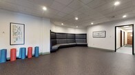 This single-floor space is designed as a versatile, multipurpose studio suited for fitness, instruction, or group activities. The main area is open and expansive, featuring durable speckled epoxy-style flooring that provides both resilience and a clean, modern aesthetic. A suspended acoustic tile ceiling with recessed lighting illuminates the space evenly, contributing to a functional and professional atmosphere.

One wall features a full-length mirror panel accompanied by a wall-mounted flat-screen display and mounted speakers, making the space well-equipped for instructional or media-driven programming. An adjacent wall showcases a subtle geometric-patterned surface treatment, adding a refined visual texture to the otherwise neutral interior. Framed abstract artwork is displayed throughout, lending a polished touch to the utilitarian setting.

A distinctive built-in curved banquette occupies one corner of the space, constructed with dark upholstered back panels and light gray seating. The high-backed design provides a defined rest or observation area, accommodating multiple occupants comfortably. Fitness accessories such as foam rollers are stored nearby, reinforcing the space's active-use purpose.

Doorways connect to adjacent corridors, facilitating easy access and circulation. The overall ambiance is clean, organized, and purposefully designed to support a range of group-oriented activities.