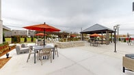 Located at 227 Pane Rd in Newington, Connecticut, this expansive outdoor amenity space is designed to offer a comprehensive al fresco living and entertaining experience. The courtyard is anchored by a striking outdoor kitchen and bar, constructed with handsome brick masonry and topped with a polished granite countertop. Bar-height seating lines the counter beneath a substantial wood-framed pergola, which is accented by pendant lantern fixtures that provide both style and evening illumination. A built-in stainless steel grill makes this an ideal space for outdoor cooking and gatherings. Across the broad concrete patio, a covered gazebo with a metal pitched roof provides a shaded retreat, surrounded by additional tables and chairs. Vibrant orange market umbrellas are distributed throughout the courtyard, adding a bold pop of color to the neutral palette of the hardscape. A raised planter with stone veneer serves as a central landscape feature, separating activity zones while adding greenery and visual interest. Elegant black lamp posts with lantern-style fixtures punctuate the space, enhancing its refined aesthetic. The perimeter is lined with neatly trimmed evergreen shrubs and enclosed by a classic black iron fence, providing both privacy and a polished, finished appearance. The multi-story building flanks one side of the courtyard, integrating seamlessly into the overall design.