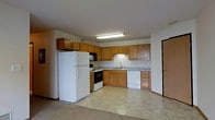 This single-floor space includes a kitchen that serves as a central functional hub, designed with practicality in mind. The kitchen features warm-toned oak cabinetry that extends to the upper walls, providing generous storage throughout. Upper and lower cabinets line the walls in an L-shaped configuration, maximizing workspace and organization. The countertops offer a neutral surface that complements the cabinetry.

Standard appliances include a full-sized white refrigerator, an electric range with oven, an over-the-range microwave, and a dishwasher, making this a fully equipped cooking space. A double sink is positioned beneath under-cabinet lighting, adding task illumination to the workspace. The kitchen floor is covered in a light-toned tile, while the adjacent area transitions to carpet, visually defining the separate zones of the space.

Overhead fluorescent lighting provides bright, even illumination throughout the kitchen. The open layout between the kitchen and the neighboring area creates a connected, accessible flow. A doorway is visible at the edge of the space, indicating a clear and practical transition to other areas of the unit.