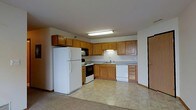 This single-floor space includes a kitchen that serves as a central functional hub, designed with practicality in mind. The kitchen features warm-toned oak cabinetry that extends to the upper walls, providing generous storage throughout. Upper and lower cabinets line the walls in an L-shaped configuration, maximizing workspace and organization. The countertops offer a neutral surface that complements the cabinetry.

Standard appliances include a full-sized white refrigerator, an electric range with oven, an over-the-range microwave, and a dishwasher, making this a fully equipped cooking space. A double sink is positioned beneath under-cabinet lighting, adding task illumination to the workspace. The kitchen floor is covered in a light-toned tile, while the adjacent area transitions to carpet, visually defining the separate zones of the space.

Overhead fluorescent lighting provides bright, even illumination throughout the kitchen. The open layout between the kitchen and the neighboring area creates a connected, accessible flow. A doorway is visible at the edge of the space, indicating a clear and practical transition to other areas of the unit.
