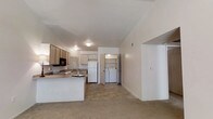 This single-floor residence offers a straightforward and functional layout designed for everyday comfort and convenience. The heart of the home is an open-concept space where the kitchen flows seamlessly into the living area, creating a cohesive and airy feel. The kitchen is equipped with light wood cabinetry, a built-in microwave, a white refrigerator, and a peninsula countertop that provides additional workspace and serves as a natural divider between the cooking and living zones. The flooring transitions from tile in the kitchen area to carpet in the living space, adding a touch of warmth underfoot. Ceiling-mounted light fixtures provide consistent illumination throughout. A standout feature of this residence is the dedicated in-unit laundry area, conveniently located just off the kitchen and partially enclosed for privacy. This space houses a full-sized washer and dryer, adding significant practical value. The hallway leading further into the unit offers access to additional rooms, with vaulted or angled ceiling lines adding subtle architectural interest. The overall ambiance is clean, neutral, and move-in ready, providing a versatile canvas for personalization.