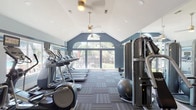This fitness center is thoughtfully designed to support a comprehensive workout experience within a bright, welcoming environment. The space is defined by its striking vaulted ceilings, which rise to an impressive peak and lend a sense of openness throughout. Arched transom windows and expansive floor-to-ceiling glass panels line the walls, flooding the interior with natural light while framing views of the surrounding greenery and outdoor areas.

The main workout floor is finished with durable, patterned carpet tiles in a coordinated gray palette, complementing the cool blue and white color scheme of the walls. The cardio zone features multiple treadmills and elliptical cross-trainers, many equipped with individual monitors. A dedicated strength training area includes a multi-station cable and weight machine, a rowing machine, free weights, and stability balls, providing a well-rounded selection of equipment for users of all fitness levels.

Full-length mirrors along one wall visually expand the space and assist with form during exercise. Pendant lighting and ceiling fans add both ambiance and ventilation. The overall atmosphere is clean, modern, and energizing, making this fitness center a functional and inviting facility for daily use.