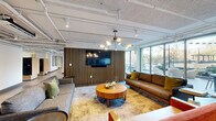 This single-floor amenity lounge is a thoughtfully designed communal space that blends social functionality with modern interior design. The open layout accommodates several distinct zones, each contributing to a versatile and inviting atmosphere. A striking slatted wood accent wall serves as the visual centerpiece of the space, framing a wall-mounted flat-screen television and anchoring the primary lounge area. Two generously sized sofas and accent seating are arranged around a round wood coffee table, creating a comfortable gathering spot ideal for relaxed conversation or media viewing. A sculptural, multi-arm chandelier adds a refined focal point overhead.

Adjacent to the lounge seating, a full-sized billiard table rests atop layered area rugs, providing an engaging recreational feature. Bar-height counter seating lines a wall near the windows, offering an informal perch with views to an outdoor terrace. Floor-to-ceiling windows flood the interior with abundant natural light and provide direct visual connectivity to the exterior patio space.

The ceiling features exposed ductwork, white-painted surfaces, and track lighting, lending an industrial-modern aesthetic throughout. Polished concrete-style flooring ties the space together, while curated artwork and decorative accents add personality and warmth to this well-appointed communal environment.