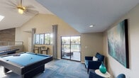 This well-appointed recreational facility offers an engaging and social atmosphere across a single thoughtfully designed floor. The centerpiece is a spacious game room featuring a full-size pool table and a shuffleboard table, set atop patterned carpet tile flooring beneath soaring vaulted ceilings. Track lighting and skylights flood the area with ample illumination, while an open mezzanine-style railing provides visual connectivity between levels of the space.

The main lounge area is an impressive, open-plan gathering space with dramatically high ceilings, multiple skylights, and abundant natural light streaming through large windows dressed with flowing curtains. A ceiling fan, pendant lighting, and track fixtures complement the bright, airy feel. The lounge is furnished with comfortable seating arrangements around a central coffee table, set against warm hardwood-style flooring and accented by a wood-paneled feature wall with a mounted television.

A cozy seating nook off the game room offers a pair of plush blue armchairs, a side table, and a large teal abstract canvas, creating an intimate retreat within the broader space. Sliding glass doors open directly onto an outdoor deck with wrought-iron railing, offering views of a pool and lush surrounding greenery — seamlessly blending indoor comfort with outdoor leisure.