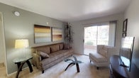 This single-floor residence offers a thoughtfully arranged living space with a warm, neutral aesthetic throughout. The living room serves as the centerpiece, finished with plush wall-to-wall carpeting in a light tone that complements the soft gray walls. A large sliding glass door floods the room with natural light and provides direct access to a private patio, creating a seamless indoor-outdoor connection. The patio overlooks a well-maintained communal landscape featuring mature palm trees, manicured greenery, and a pool area visible through decorative iron fencing — offering a pleasant and tranquil outdoor view.

Inside the living room, the layout comfortably accommodates a full sofa, accent chair, and a sculptural glass-top coffee table, all anchored beneath abstract wall art that adds a contemporary touch. A tall indoor plant in the corner adds a natural element to the space. The overall ambiance is calm and inviting, with an emphasis on natural light and open sightlines to the outdoors, making this single-story space feel airy and connected to its surroundings.