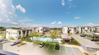 This contemporary residential community presents a well-appointed, Florida-inspired living environment spread across a thoughtfully designed campus. The exterior showcases modern multi-story buildings finished in clean, neutral tones with structured facades that reflect a consistent and polished architectural style throughout the property. At the heart of the community lies a resort-style pool area, complete with lounge seating, umbrellas, and teal-blue waters, all framed by lush tropical palm trees that lend a distinctly warm-weather ambiance to the surroundings. The landscaping is carefully curated, with greenery and palms providing both shade and visual appeal around the pool deck. Ample surface parking is conveniently distributed throughout the property, ensuring accessibility for residents and guests alike. The elevated vantage point of certain areas within the community offers sweeping views of the pool area, neighboring buildings, and the open sky beyond — a feature that enhances the overall sense of space and openness. The community's layout promotes a connected, walkable environment with clear sightlines across common areas. The combination of modern architecture, resort amenities, and tropical landscaping creates an inviting and vibrant living atmosphere well-suited to a sunny, warm climate.