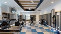 The fitness center at 1319 Fonthill Court in Langhorne, PA offers a thoughtfully designed workout facility that balances functionality with contemporary style. Guests are welcomed through a bright, airy entry foyer featuring stone-tile flooring, soaring ceilings, arched doorways, and large glass entry doors that allow generous natural light to fill the space.

The main exercise area is defined by a striking coffered ceiling with exposed rustic wood beams, a ceiling fan, and recessed lighting that creates a warm yet energizing atmosphere. The walls feature a distinctive dark vertical-slat paneling with wood trim accents, lending a modern, upscale aesthetic throughout. A bold blue-and-tan checkered carpet tile floor anchors the space and adds visual interest.

Cardio equipment, including treadmills, ellipticals, and a stationary bike, is positioned near large windows that overlook outdoor greenery, providing natural light and pleasant views during workouts. A wall-mounted flat-screen television adds entertainment options for cardio users.

The strength training area includes a weight bench, cable machine, and a well-organized dumbbell rack. A dedicated functional training alcove houses resistance bands, stability balls, a BOSU trainer, and medicine balls, offering a comprehensive range of workout options within this efficiently organized and attractively appointed fitness facility.