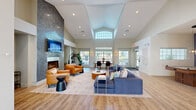 Located at 100 Christina Mill Dr in Newark, Delaware, this single-floor community amenity space offers a thoughtfully designed layout across a generous footprint. The centerpiece is a dramatic open lounge area distinguished by soaring vaulted ceilings and an abundance of natural light streaming through large transom windows. A floor-to-ceiling textured stone fireplace wall anchors the room, complemented by a wall-mounted television and recessed lighting throughout. The space is furnished with a sectional sofa, leather accent chairs, a round coffee table, and an area rug, creating a warm and inviting atmosphere. Light wood-style flooring runs throughout, adding continuity and warmth. Adjacent to the main lounge is a dining area with a round wood table and modern chairs, ideal for casual gatherings. A bright, airy hallway with a chandelier and glass-panel entry doors connects the various areas seamlessly. A dedicated package room houses multiple banks of smart lockers, including Amazon Hub lockers, providing a secure and convenient package management solution. An additional flexible room features modern track lighting, curated wall art, and understated contemporary furnishings, suitable for a variety of uses. The overall ambiance is modern, clean, and welcoming.