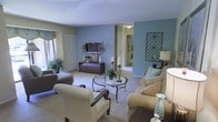 This single-floor residence offers a thoughtfully designed interior that balances comfort and style. The living room serves as the heart of the space, showcasing a striking blue accent wall that adds depth and character to the room. Plush, neutral-toned carpeting runs throughout, contributing to the warm and inviting ambiance. The seating arrangement includes a generously sized sofa, a classic checkered armchair, and a chaise lounge, all oriented around a elegant round glass-top coffee table with a wrought iron base. A wall-mounted flat-screen television sits above a rich dark-wood media console, which anchors the entertainment area. Decorative elements such as candle holders, potted greenery, and wall art add personality and visual interest throughout the space. Natural light filters in through a window dressed with a decorative valance, brightening the room and complementing the soft overhead lighting. A hallway visible from the living area leads to additional rooms beyond, suggesting a well-connected and functional floor plan. The overall aesthetic is cohesive and polished, with careful attention to detail in both color palette and furnishing choices, making this a welcoming and livable space.