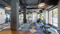 The fitness center at 255 Huguenot St in New Rochelle, NY is a thoughtfully designed, single-floor facility that combines industrial aesthetics with a comprehensive range of workout options. The space features exposed ceilings with visible ductwork and structural concrete columns, complemented by stylish drum pendant lighting that adds a refined touch to the raw architectural framework. Large windows along the perimeter flood the interior with natural light and provide views of the surrounding streetscape.

The main workout floor is finished with a distinctive blue and tan checkered carpet tile, creating defined zones throughout. A robust cardio section lines the window wall, equipped with TRUE treadmills, ellipticals, and Cybex cycling bikes. Rowing machines and cable machines are distributed throughout the open floor plan, alongside free weight stations with dumbbell racks and a dual water fountain set against a decorative faux-greenery accent wall.

A dedicated functional training area features rubber flooring in contrasting gray and green tones, housing power racks with barbells, a heavy bag, TRX suspension trainers, kettlebells, medicine balls, and a tire sled—catering to high-intensity and strength-focused workouts. The overall ambiance is energetic and modern, well-suited for a wide range of fitness pursuits.